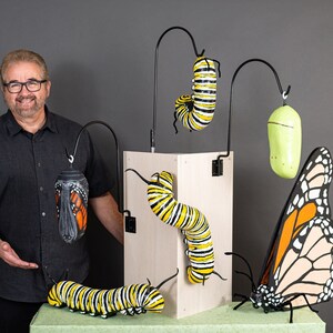 May include: A man stands in front of a display of monarch butterfly life cycle sculptures. The sculptures include a caterpillar, chrysalis, and butterfly, all painted in realistic detail. The sculptures are displayed on a wooden stand.