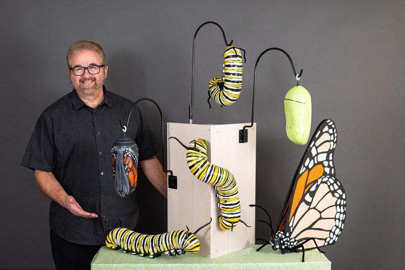 May include: A man stands in front of a display of monarch butterfly life cycle sculptures. The sculptures include a caterpillar, chrysalis, and butterfly, all painted in realistic detail. The sculptures are displayed on a wooden stand.
