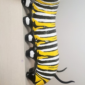 May include: A black and yellow striped caterpillar sculpture with white spots. The caterpillar is painted with a glossy finish and has two black antennae.