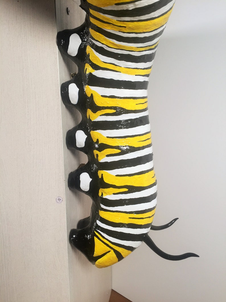 May include: A black and yellow striped caterpillar sculpture with white spots. The caterpillar is painted with a glossy finish and has two black antennae.