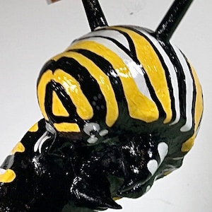 May include: Close-up of a black and yellow striped ceramic sculpture of a caterpillar. The caterpillar has a white stripe on its head and black antennae.