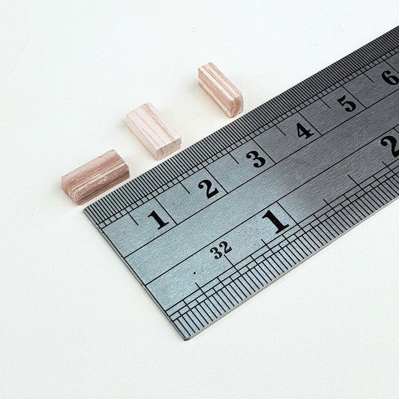 Może przedstawiać: Trzy drewniane klocki o r&oacute;żnych rozmiarach umieszczono na metalowej linijce. Linijka jest oznaczona liczbami od 1 do 6. Klocki są wykonane z tego samego rodzaju drewna, kt&oacute;re ma jasnobrązowy kolor.