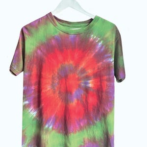 May include: A tie-dye t-shirt in a spiral pattern, featuring a blend of red, purple, and green hues. The shirt is hanging on a white hanger against a white background. The short-sleeved tee is a casual wear item.