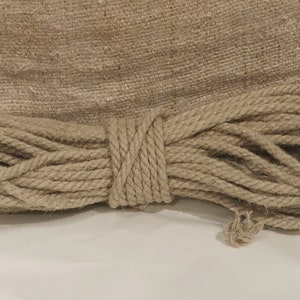 Peut inclure: Une corde en fibre naturelle enroulée, probablement en jute ou en chanvre, avec une section centrale bien serrée. La corde est de couleur marron clair et semble neuve. Un morceau de tissu de jute est en arrière-plan.