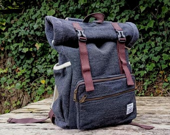 May include: A dark gray canvas backpack with brown straps and a roll-top closure. The backpack has a zippered pocket on the front and a small patch with a logo on the bottom right corner.