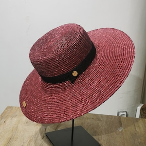 Op de afbeelding: Een brede strohoed met een zwart lint en een klein bruin label. De hoed is diep roze van kleur met een glinsterende afwerking.