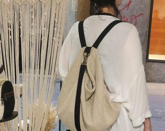 Könnte beinhalten: Ein beigefarbener Canvas-Rucksack mit schwarzen Trägern. Der Rucksack hat einen lässigen, schlabberigen Look und wird von einer Person getragen.
