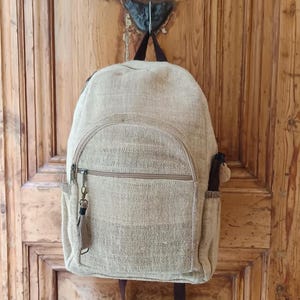 Op de afbeelding: Een beige rugzak van een geweven stof, hangend aan een houten deur. De rugzak heeft een hoofdvak, een voorvak met rits en zijvakken. Het heeft bruine ritsen en bandjes, met een kleine sleutelhanger aan de rits.