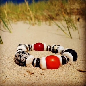 Könnte beinhalten: Ein Armband aus weißen, schwarzen und roten Perlen. Die Perlen sind in einem Muster angeordnet und auf einer Schnur aufgefädelt. Das Armband liegt auf einem Sandstrand.