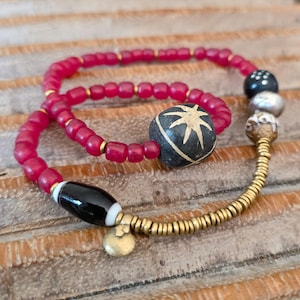 Puede incluir: Una pulsera hecha a mano con una mezcla de cuentas de varias formas y colores. La pulsera incluye pequeñas cuentas rojas redondas, espaciadores dorados y cuentas decorativas más grandes en negro, blanco y plateado. Un pequeño dije dorado cuelga de la pulsera.
