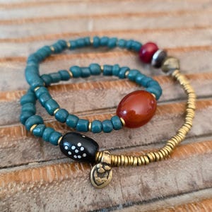 Puede incluir: Dos pulseras de cuentas. Una pulsera presenta cuentas de color turquesa con detalles dorados y una cuenta grande, ovalada, de color marrón rojizo. La otra pulsera tiene cuentas doradas y una cuenta negra con puntos blancos.