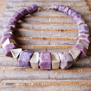 May include: A necklace with large, irregular, purple gemstones and silver-coloured metal pieces. The necklace is displayed on a textured, light brown surface, highlighting its unique design and colour contrast. The necklace is approximately 46 cm in length.