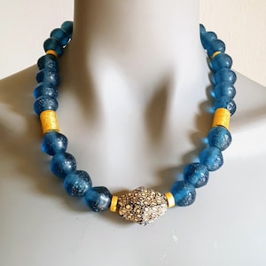 Könnte beinhalten: Halskette mit großen, runden, durchscheinenden blauen Perlen, die von goldfarbenen Zylinderperlen unterbrochen werden. Der Mittelpunkt ist eine gemusterte, goldfarbene Perle. Die Halskette wird auf einer Schaufensterpuppe präsentiert.