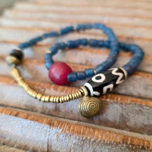 Könnte beinhalten: Zwei Armbänder mit Perlen. Ein Armband hat blaue Perlen und eine rote Perle. Das andere Armband hat blaue Perlen, eine schwarz-weiß gemusterte Perle und goldfarbene Metallakzente. Die Armbänder liegen auf einer strukturierten Oberfläche.