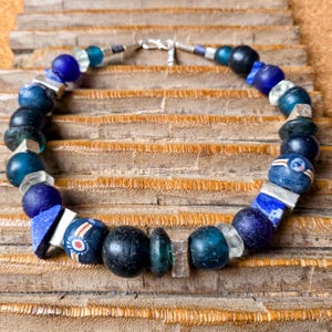 May include: A handcrafted necklace with a mix of blue glass beads, including round, square, and faceted shapes. The beads are separated by silver-toned metal accents and a silver clasp. The necklace is presented on a wooden surface.