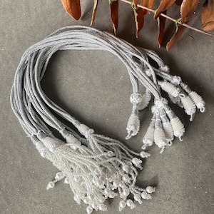 May include: A bundle of silver and white beaded tassels with a silver cord. The tassels are made of small white beads and have a silver cord at the top.
