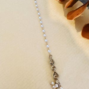 Op de afbeelding: Een goudkleurig hoofddeksel met een delicaat ontwerp. Een ketting van kleine witte parels leidt naar een decoratief gedeelte met druppelvormige accenten en kleine, ronde en peervormige parels. Het hoofddeksel is ontworpen voor speciale gelegenheden.