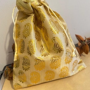 May include: A gold drawstring bag featuring a paisley pattern in a lighter gold hue. The bag is crafted from a shiny material and has a matching gold drawstring closure. The bag rests on a light brown surface.