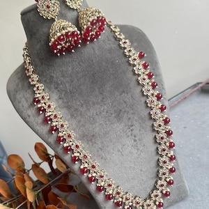 Könnte beinhalten: Ein goldenes Halsketten- und Ohrring-Set mit roten Perlen und floralen Akzenten. Die Halskette ist lang und hat ein zartes Design. Die Ohrringe sind baumelnd mit einem glockenförmigen Anhänger.