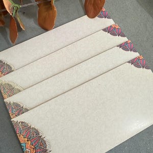 May include: A collection of cream-coloured envelopes with a subtle floral pattern. Each envelope corner displays a vibrant, ornate design in red, blue, orange, and gold. The envelopes are arranged on a grey surface.