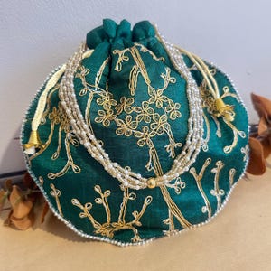 May include: A round, emerald green drawstring pouch with gold floral embroidery and a beaded strap. The pouch has a white beaded trim and a gold tassel. The bag is approximately 15 cm in diameter.