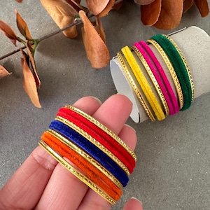 Op de afbeelding: Een set van zes gouden armbanden met fluweel in rood, oranje, blauw, geel, roze en groen.