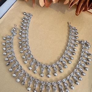 May include: A pair of silver-toned anklets featuring a series of oval-shaped clear stones set in metal frames. Each stone is accented by a small, round, dangling silver-toned bell charm attached to the bottom, creating a rhythmic, layered appearance.