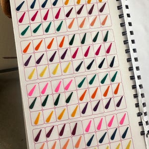 Puede incluir: Una página de cuaderno en espiral con pegatinas kumkum en forma de lágrima de varios colores, incluyendo rosa, verde, naranja, amarillo y morado. El texto "A QUALITY PRODUCT" y "STICKER KUMKUM" es visible.