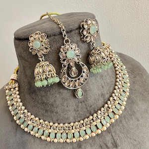 Pode incluir: Um conjunto de joias composto por um colar, dois brincos jhumka e um maang tikka. As peças são feitas de metal dourado com pedras ovais verde-menta claro, cristais transparentes e pequenas pérolas brancas em padrões florais intrincados.