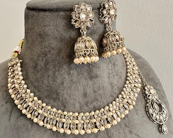 Könnte beinhalten: Ein goldfarbenes Schmuckset bestehend aus einer Halskette und passenden Jhumka-Ohrringen. Die Halskette ist mit Reihen von klaren Steinen im Marquise-Schliff und kleinen cremefarbenen Perlen verziert. Die glockenförmigen Ohrringe haben florale Aufsätze.