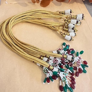 May include: A set of gold metallic cords featuring decorative tassels at the ends. The tassels are adorned with white seed beads, clear spherical beads, and dangling oval-shaped beads in deep red and emerald green colours, secured with gold thread.