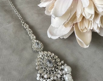 May include: A silver and white bridal tikka with a large, ornate, crystal-encrusted pendant and a chain of smaller crystals. The pendant is surrounded by a fringe of white pearls.