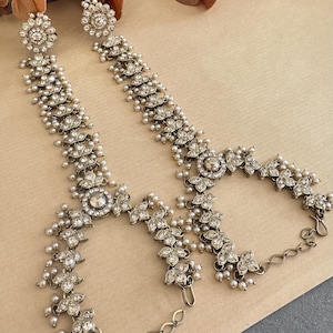 May include: Silver headpiece with floral and pearl details. The design incorporates flower-shaped elements, each set with clear crystals. Tiny pearls are placed throughout, creating a delicate and elegant appearance. The headpiece is a beautiful accessory.