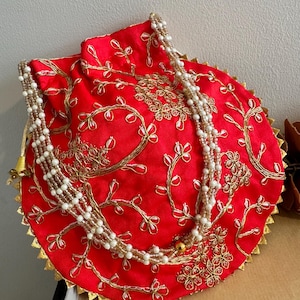 Puede incluir: Un bolso redondo rojo con bordados dorados y una correa de cuentas. El bolso presenta intrincados patrones florales y un ribete dorado en el borde. La correa está hecha de cuentas blancas y doradas.