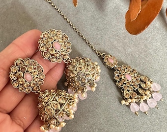 May include: A set of gold-tone earrings and a matching tikka, all adorned with sparkling rhinestones and pink accents. The earrings feature a dangling design with a round top and a bell-shaped bottom.