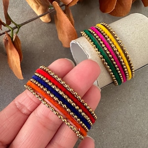 May include: Set of six colorful velvet bangles with gold bead accents. The bangles are stacked in a rainbow pattern of orange, red, blue, green, pink, and yellow.