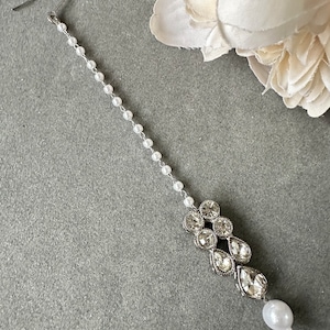 May include: A silver and white pearl maang tikka with a teardrop-shaped pearl at the end. The maang tikka is made of a chain of small white pearls and a silver pendant with clear rhinestones.