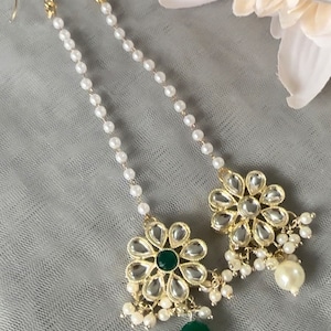 May include: A pair of gold earrings with a pearl chain and a floral design. The floral design features a green gem in the center and is surrounded by clear rhinestones. The earrings are accented with small pearl drops.