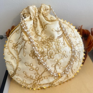Peut inclure: Sac à cordon de serrage crème avec broderie florale dorée et une bandoulière perlée. Le sac a une bordure dorée festonnée et une fermeture à cordon dorée. La bandoulière perlée est blanche et dorée. Le sac est rond.
