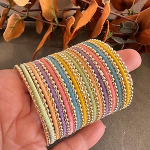 May include: A stack of multiple round bracelets featuring alternating bands of pastel-coloured velvet and metallic gold-toned beads. The colours include light green, soft orange, lavender, pink, blue, and yellow, arranged in a repeating pattern.