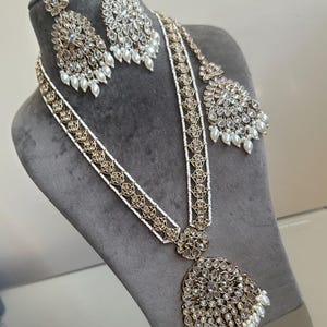 May include: A gold and white layered necklace set with a large pendant and matching earrings. The necklace features a delicate chain with small white pearls and a larger chain with gold accents and clear stones. The pendant is a large, ornate design with clear stones and a pearl drop. The earrings are similar in design to the pendant, with a smaller version of the pendant and a pearl drop.