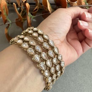 Op de afbeelding: Goudkleurige armband met meerdere rijen heldere, druppelvormige stenen. De armband wordt om een pols gedragen, met een neutrale achtergrond en gedroogde bladeren in het bovenste deel van de afbeelding. Een opvallend sieraad.