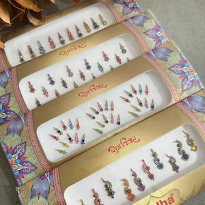 May include: Four boxes of Sudha Fancy Bindi, each with numerous decorative forehead jewels. The bindis are in various colours, including red, green, blue, and gold. The boxes have gold and purple floral designs.