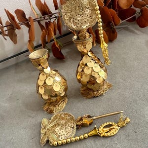 May include: Gold decorative tassels with sequins and beaded detailing. The tassels feature a bulbous form, embellished with gold thread and small, round sequins. One tassel lies flat, while two others stand upright. A sprig of brown leaves is in the background.