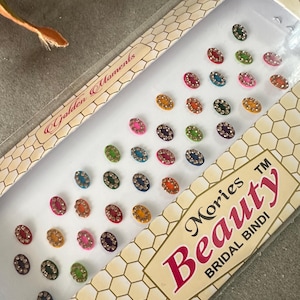 Op de afbeelding: Een doos met kleurrijke ovale bindis met juwelen. De doos is wit met een gouden honingraatpatroon en de tekst "Golden Moments Mories TM Beauty Bridal Bindi" in rood en zwart.