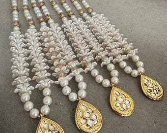 May include: Several strands of white pearl necklaces with gold-colored teardrop pendants. The pendants have a decorative, openwork design. The necklaces are arranged on a neutral surface, with some decorative foliage in the background.