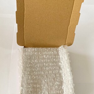 Może przedstawiać: Brązowe pudełko kartonowe z otwartą klapą, ukazujące przezroczystą folię bąbelkową z plastiku w środku.
