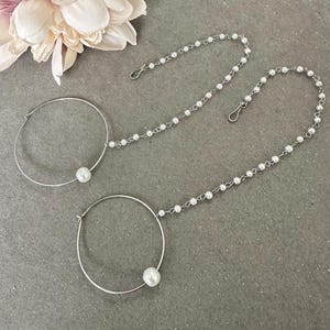 May include: Two silver nose rings with a pearl attached to each ring. Each ring is connected to a silver chain with white pearls.