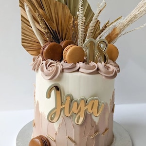 Puede incluir: Un pastel blanco con glaseado marrón y beige decorado con hojas de palma secas, hierba de la pampa y macarons. El pastel está cubierto con purpurina dorada y el número 18 en dorado. El nombre "Jiya" está escrito en dorado en el lateral del pastel.