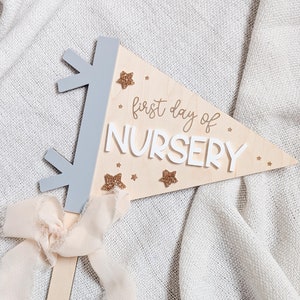 May include: A wooden pennant with the text "first day of NURSERY" in white letters. The pennant is decorated with gold glitter stars and has a light blue wooden base.